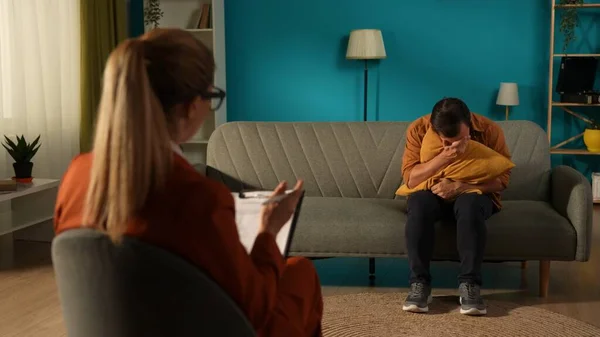 A female psychologist working with an unhappy man suffering from depression. A man is sitting on a couch in front of a specialist, hugging a pillow. Concept of mental health, depression, panic attacks