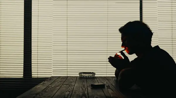 シルエットショットで 若い男がカフェのテーブルに座っている テーブルにはタバコと灰皿があります タバコに煙を灯している 悲しみを示しています ミディアムショット — ストック写真