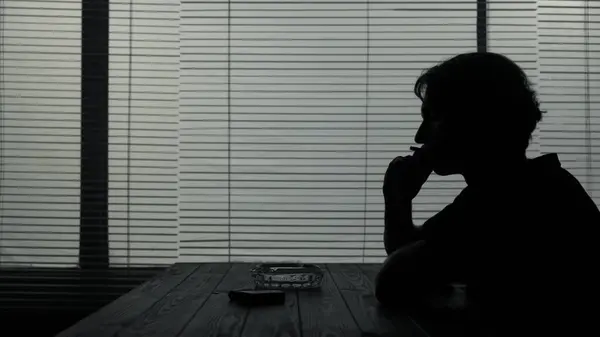 シルエットショットで 若い男がカフェのテーブルに座っている テーブルの上にはタバコと灰皿があります タバコを吸ってどこかで見ている 悲しみを示す — ストック写真