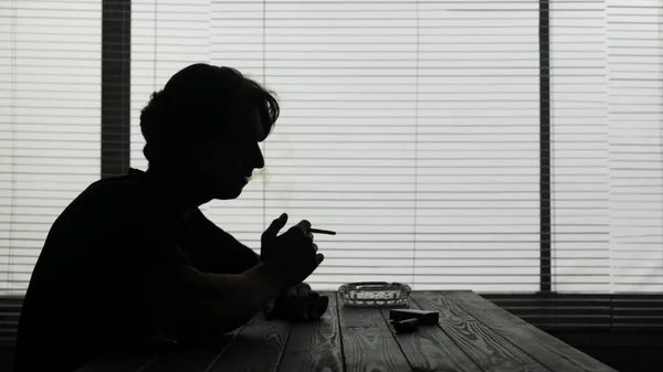 カフェのテーブルに座っている若者のシルエット 彼はタバコを吸い 煙を放ち どこかで見ている 悲しみを示しています ミディアムショット — ストック写真