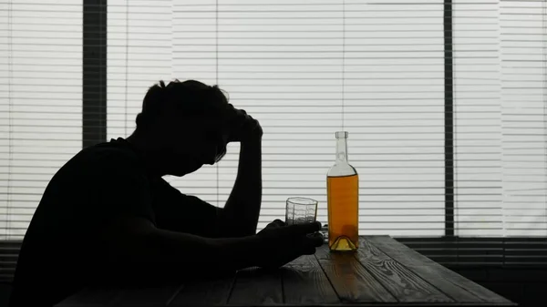 カフェのテーブルに座っている若者のシルエット 近くには 彼の手にアルコールのボトルがあり 彼はグラスを持っており 何かを考えている アルコール依存症 悲しみ 孤独を示しています — ストック写真
