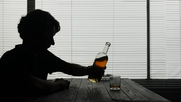 In the frame, a young man is sitting in a cafe. He is frustrated, sad. Then takes a bottle pours it into a glass and drinks the alcohol. Demonstrates addiction to alcohol, loneliness, sadness.