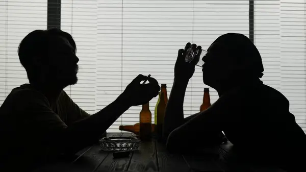 若い人々は施設のテーブルに座っている 空のボトルに囲まれている タバコを点灯する もう一人はグラスから飲み物を終える エイリアンは何かについて話す ミディアムショット — ストック写真
