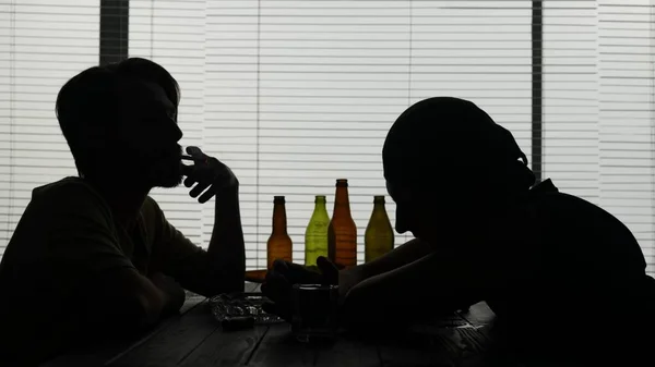 若い人々は施設のテーブルに座っている 空のボトルに囲まれている タバコを点灯する もう一人はグラスから飲み物を終える エイリアンは何かについて話す ミディアムショット — ストック写真