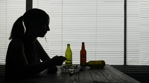 フレームには 若い女性が空のボトルに囲まれたテーブルの上に座っています 彼女はアルコール中毒を示している 彼女はタバコを吸って それを終えた後 それを取り出した 孤独なシェイズ 悲しい — ストック写真