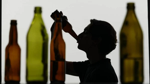 A close up of multicolored empty bottles on a white background. In the background is a silhouette of a man. He sits sideways to the camera and drinks alcohol from the bottle. Medium shot.