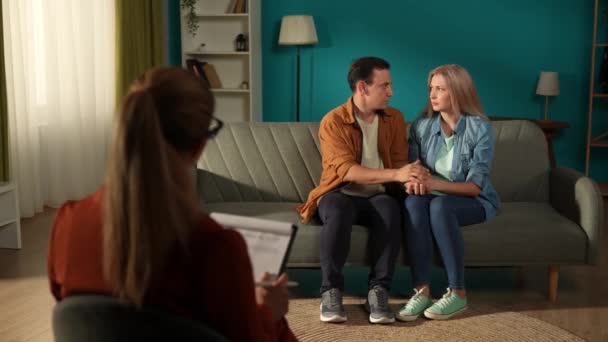 家族問題の専門家とのレセプションで結婚したカップル 男と女が手を握り ソファーに座りながら問題を共有する セラピストは彼らに質問し メモを取る — ストック動画