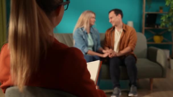 ファミリーカウンセリング 女性心理学者の前でソファーに座っている夫婦の手を握る 心理療法セッション中にノートブックにメモを取る女性心理学者 — ストック動画