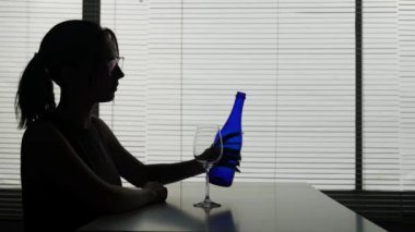 Genç bir kadın bir masada, yanında alkol ve cam olan bir şişe de yanında. Biraz dökülüyor, bardakları ve içkileri inceliyor. Alkolik bağımlılığı, yalnızlığı ve üzüntüyü gösterir..
