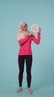 Tam boy, izole bir fotoğraf spor giyimli ve tesettürlü çekici bir genç kadın, Sheila. O bir saat tutuyor ve gülümsüyor. Reklam için yer, promosyon, spor.