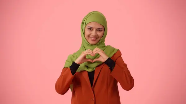 Medium-sized isolated photo capturing an attractive young woman wearing a hijab, veil. She is making a heart with her hands and smiling. Place for your advertisement, cultural diversity.