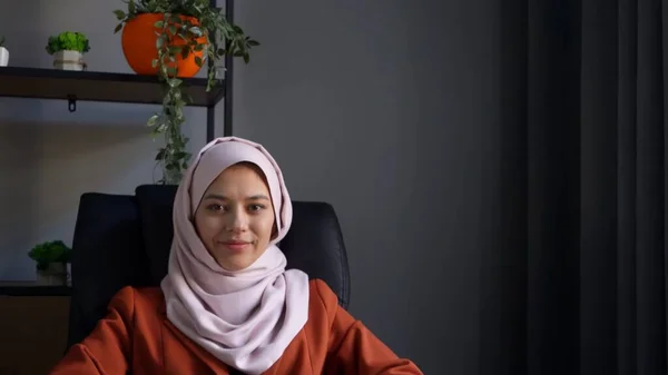 Medium-sized photo capturing an attractive young woman wearing a hijab, veil. She is sitting in the office, leaning back in the chair and smiling. Cultural diversity, job and education, advertisement.