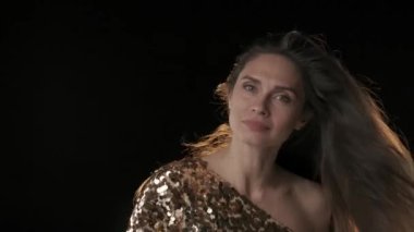 Siyah arka planda orta yaşlı, siyah saçlı bir kadın var. Kameraya bakıyor, başını sallıyor ve saçlarını sallıyor. Rüzgar ona esiyor ve saçlarını büyütüyor. Ağır çekim. HDR BT2020 HLG