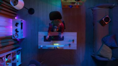 Çevrimiçi oyun reklam konsepti. Evde modern bilgisayar olan bir oda manzarası. Kulaklık takmış bir kızın masada oturup bilgisayar başında oyun oynaması..