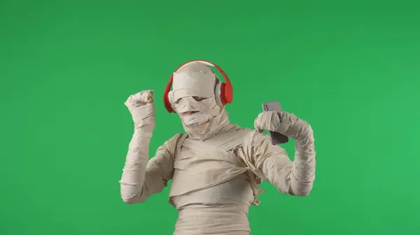 Green screen isolated chroma key photo capturing a mummy wearing headphones and dancing to the music with a smartphone in its hand. Halloween holidays. Mock up for your promotion clip or advertisement