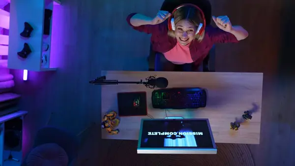 Gaming and streaming advertisement concept. Person on the PC at home. Top view of woman in headphones at the computer, won the mission in the game, looking up holding hands up.