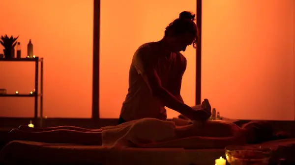 Masseur, massage specialist giving a massage to his patient using herbal bags. Silhouettes in the massaging room, spa procedure. Orange neon lights. Healthcare, medical treatment, holistic therapy.