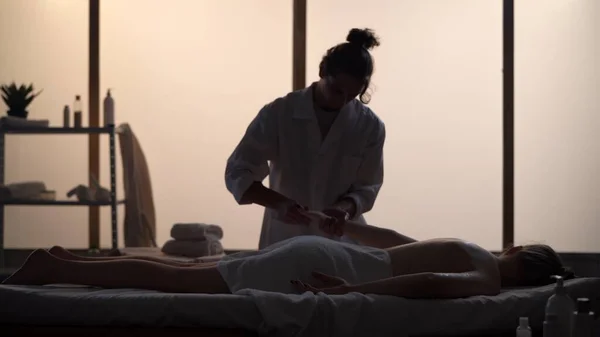Masseur, massage specialist giving back and arm massage to his patient. Silhouettes of a woman and a man in the massaging room, spa procedure. Healthcare, medical treatment, holistic therapy.