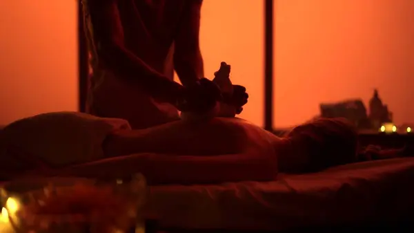 Masseur, massage specialist giving a massage to his patient using herbal bags. Silhouettes in the massaging room, spa procedure. Orange neon lights. Healthcare, medical treatment, holistic therapy.