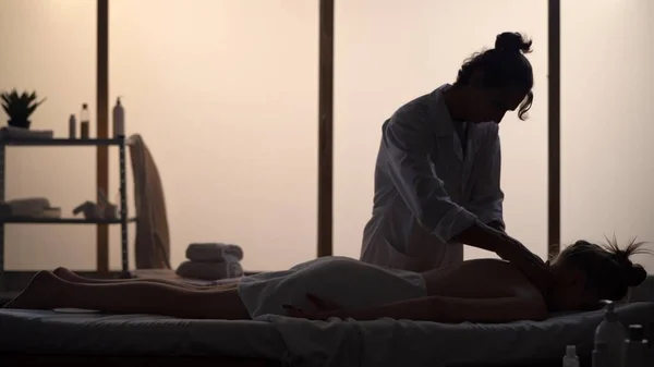 Masseur, massage specialist giving neck massage to his patient. Silhouettes of a woman and a man in the massaging room, spa procedure. Healthcare, medical treatment, holistic therapy.