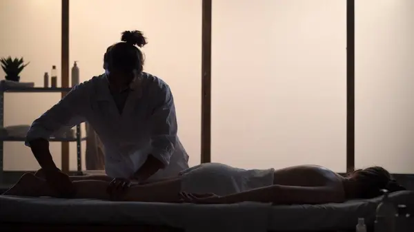 Masseur, massage specialist giving full-body massage to his patient. Silhouettes of a woman and a man in the massaging room, spa procedure. Healthcare, medical treatment, holistic therapy.