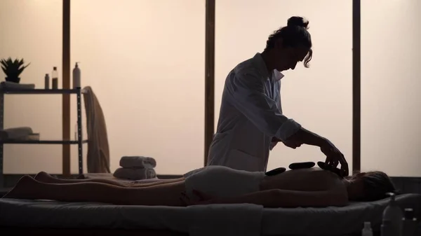 Masseur, massage specialist giving a relaxing massage using heated rocks. Silhouettes of a woman and a man in the massaging room, spa procedure. Healthcare, medical treatment, holistic therapy.