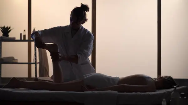Masseur, massage specialist giving a foot and leg massage to his patient. Silhouettes in the massaging room, spa procedure. Healthcare, medical treatment, holistic therapy.
