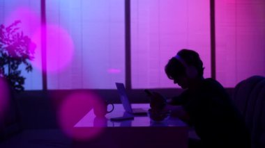 Günlük yaşam sosyal ilişkiler siluet konsepti. Kafeteryadaki erkek portresi. Gece fast food barında oturan, kulaklıklı, akıllı telefondan müzik dinleyen, laptopunu masaya koyan bir adam..