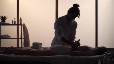 Masör, masaj uzmanı, bitkisel poşetler kullanarak hastasına masaj yapıyor. Masaj odasında bir kadınla bir erkeğin siluetleri, spa prosedürü. Sağlık, tıbbi tedavi, bütünsel terapi.