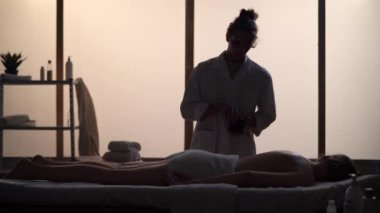 Masör, masaj uzmanı ısıtmalı taşlarla rahatlatıcı masaj yapıyor. Masaj odasında bir kadınla bir erkeğin siluetleri, spa prosedürü. Sağlık, tıbbi tedavi, bütünsel terapi.