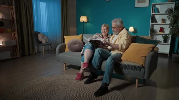 フレームには 高齢のカップルが青い壁に対してアパートのソファーに座っています 彼らは写真のアルバムを見つめている 彼らは 古い時代を思い出し ノスタルジックでチャットしています — ストック動画