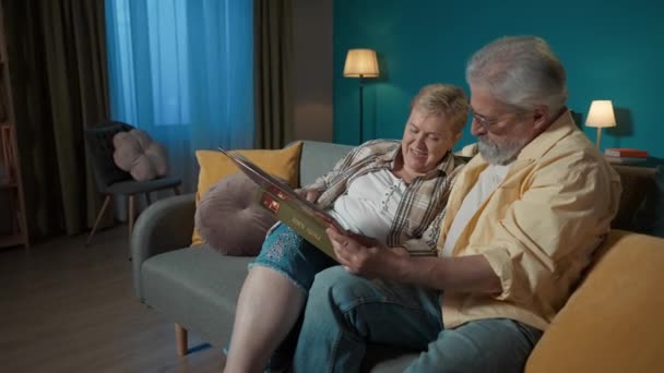 フレームには 高齢のカップルが青い壁に対してアパートのソファーに座っています 彼らは写真アルバムを見ている 彼らは 古い時代を思い出し ノスタルジックでチャットしています ミディアムビデオ — ストック動画