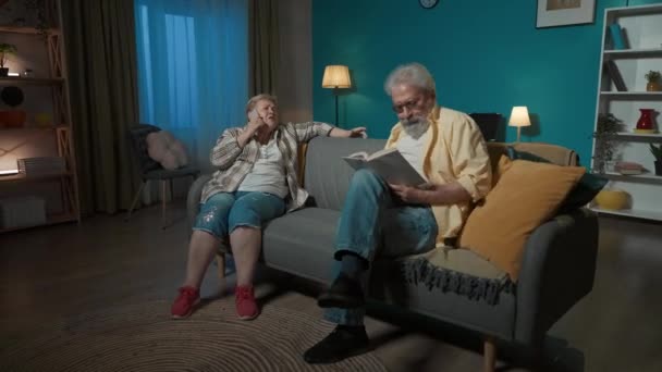 年配のカップルがソファーに座っています 女性は電話で情熱的に話している 本を読んでいる男に干渉する 彼はそれを好まない 彼は自由ではない 彼女は幸せで楽しい — ストック動画