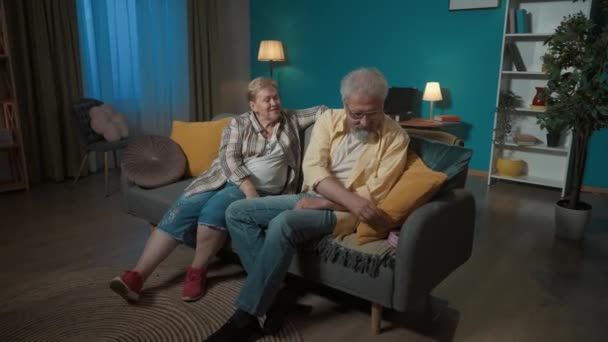 写真では 高齢のカップルは青い壁に対してアパートのソファーに座っています 男は女に贈り物をした 彼女は驚き 興奮し 喜んでいる ロマンス ミディアムビデオ — ストック動画
