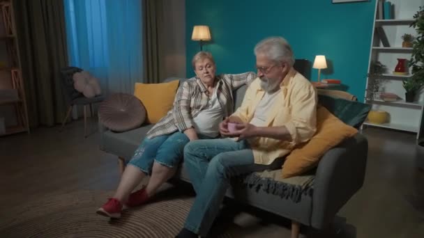写真では お年寄りのカップルが部屋のソファーに座っています 男性は枕の下に隠されたピンクの箱に手を伸ばします 女性のための贈り物は 彼女を驚かせたい 彼女は見ていないか知らない — ストック動画