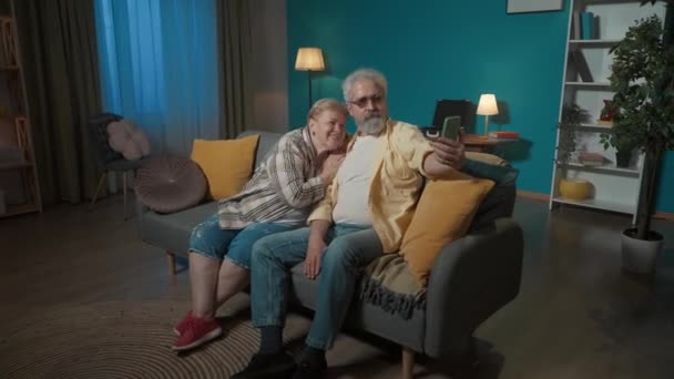 在照片中 一对老夫妻坐在公寓的沙发上 那个男人拿着电话 他们摆出一副自拍的架势 然后男人给女人看了照片 选了一个合适的 — 图库视频影像