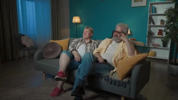 写真では 高齢のカップルがアパートのソファーに座っており 何かがテレビで面白い映画や番組を見ています 彼らは笑い 楽しむことを楽しんでいる メディアビデオ — ストック動画