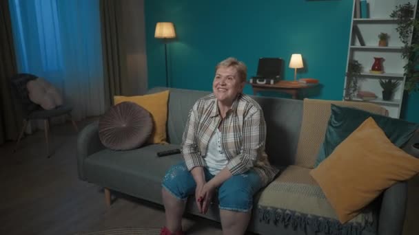 写真では 高齢の女性がアパートのソファーに座っています まるで映画や番組を見ているかのようだ 彼女は幸せで 彼女は笑っている 彼女は面白いです 中型ビデオ クローザー — ストック動画