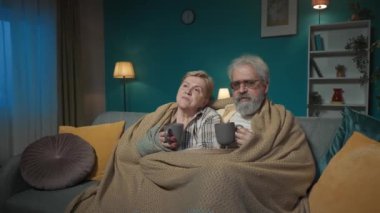 Resimde, yaşlı bir çift battaniyenin altındaki bir odada bir kanepede oturuyor. Bir fincan sıcak çay ya da kahve tutuyorlar. TV 'de bir film ya da yayın izle. Rahatlık göstergesi. Orta boy video.