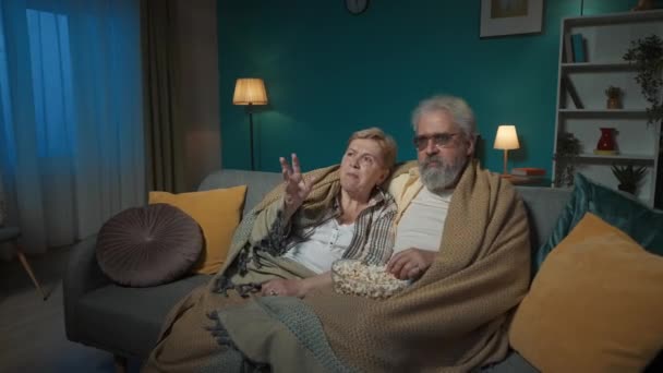 フレームには 高齢のカップルがソファーの下に座っています 彼らはテレビで映画や番組を見たり ポップコーンを食べたりしている 彼らは積極的に何かを議論し コミュニケーションしています — ストック動画