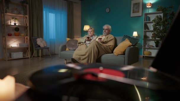 前景フレームでは ビニールターンテーブルスピンレコード 背景には 高齢のカップルがワインのグラスとソファーの下に置かれている 彼らは音楽を聴き 楽しんでいる — ストック動画