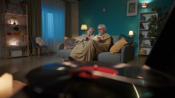 前景フレームでは ビニールターンテーブルスピンレコード 背景には 高齢のカップルがワインのグラスとソファーの下に置かれている 彼らは音楽を聴き 楽しんでいる — ストック動画
