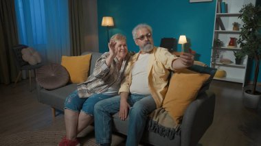 Resimde, yaşlı bir çift bir odada bir kanepede oturuyor. Adam kolunda bir telefon tutuyor. Kadın adama sarılıp yatmış ve ikisi de telefona bakmış. Fotoğraf pozunu göster.