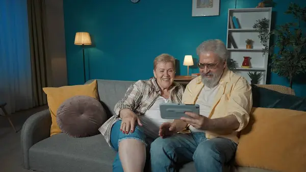 In the picture, an elderly couple is sitting on a sofa in a room. The man is holding a tablet and they are both looking at him. Demonstrate watching videos or browsing websites. They are happy.