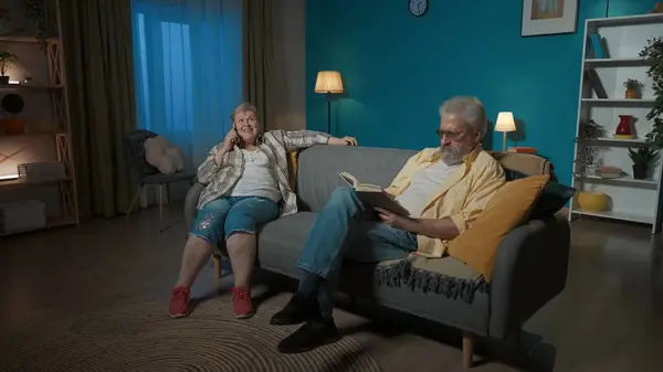 An elderly couple sits on the sofa in the room. The man reads the book carefully. The woman is passionately talking on the phone, she is happy. Apparently she is preventing a man from reading a book.