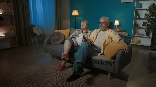 In the picture the elderly couple is sitting on the sofa in the apartment. They look aside, as in the TV. Watch a movie or show, they do not like it. Overall plan.