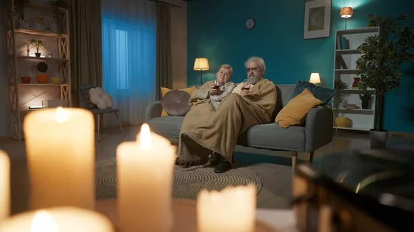 The shot shows a close-up of a vinyl record player and burning candles. In the background, an elderly couple sit under a plaid on a sofa with a glass of wine and stare intently into the camera.