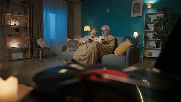 The shot shows a close-up of a vinyl record player and burning candles. In the background, an elderly couple sit under a plaid on a sofa with a glass of wine and stare intently into the camera.