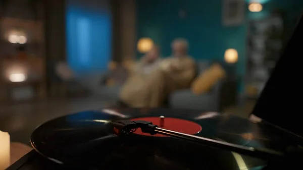 Close up shot shows a vinyl turntable spinning a record. The camera then moves to the side and burning candles of different sizes are seen. In the background are a couple of people sitting on a sofa.