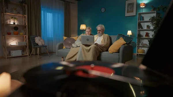 The close up picture shows a vinyl record player with a spinning disc. In the background, an elderly couple is sitting on a couch with a laptop and surfing websites and social media. Medium shot.
